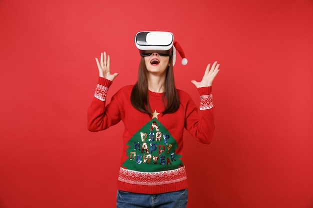 Emocionada joven Santa con sombrero de Navidad mirando hacia arriba en auriculares extendiendo las manos, manteniendo la boca abierta aislada sobre fondo rojo brillante de la pared. Feliz año nuevo 2019 celebración concepto de fiesta navideña.