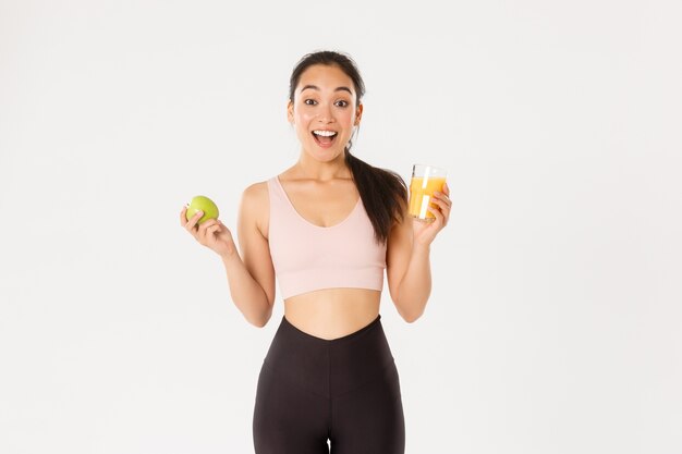 Emocionada linda chica asiática fitness, deportista con jugo de manzana y naranja jadeando sorprendida y feliz, comiendo sano para mantenerse en forma