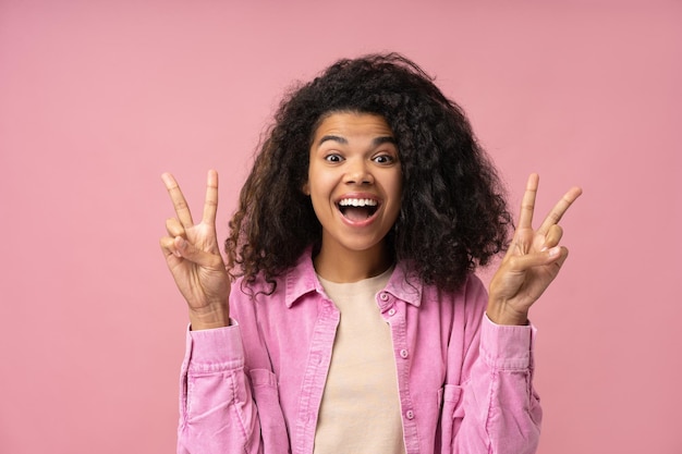 Emocionada mujer afroamericana mostrando el signo de la victoria mirando a la cámara aislada en el fondo rosa