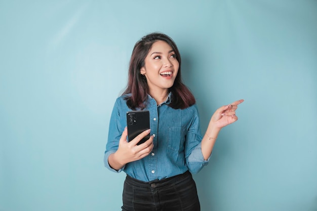 Emocionada mujer asiática con pantalones azul apuntando al espacio de copia a su lado mientras sostiene un teléfono inteligente aislado por fondo azul