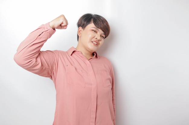 Emocionada mujer asiática de pelo corto con una camisa rosa que muestra un gesto fuerte levantando los brazos y los músculos sonriendo con orgullo