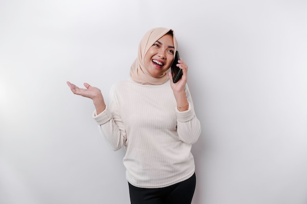 Emocionada mujer asiática usando hijab señalando el espacio de copia a su lado mientras sostiene su teléfono aislado por fondo blanco