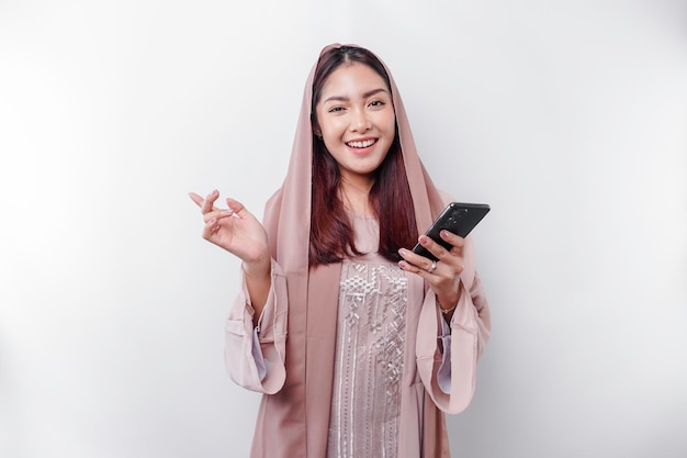 Emocionada mujer asiática usando hijab señalando el espacio de copia a su lado mientras sostiene su teléfono aislado por fondo blanco
