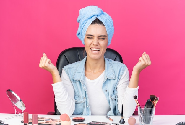 Emocionada mujer bonita caucásica con el pelo envuelto en una toalla sentado en la mesa con herramientas de maquillaje manteniendo los puños en alto