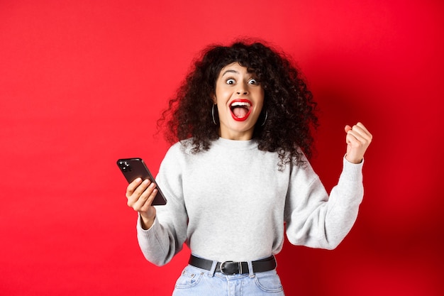 Emocionada mujer caucásica con cabello rizado, cantando, ganando un premio en línea, sosteniendo un teléfono inteligente y triunfando, de pie sobre un fondo rojo.