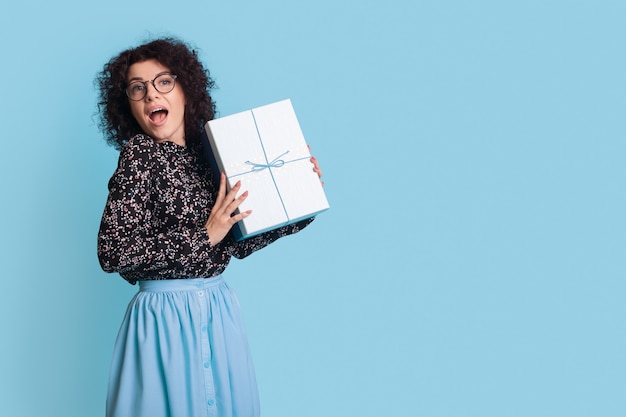 Emocionada mujer caucásica sostiene un regalo en una pared de estudio azul con espacio libre con un vestido y gafas