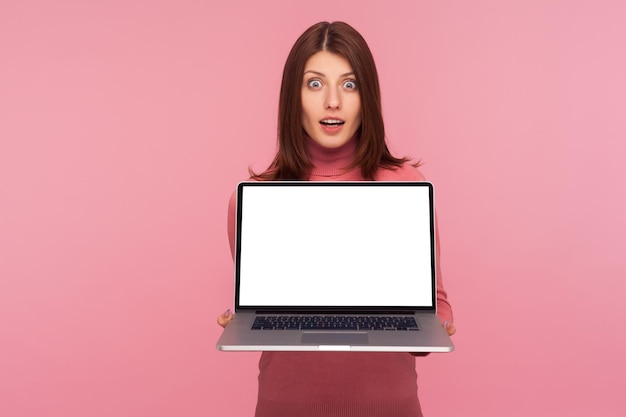 Emocionada mujer feliz asombrada con cabello castaño sosteniendo una computadora portátil con pantalla vacía que muestra una pantalla en blanco simulada para publicidad web texto comercial Estudio interior aislado en fondo rosa