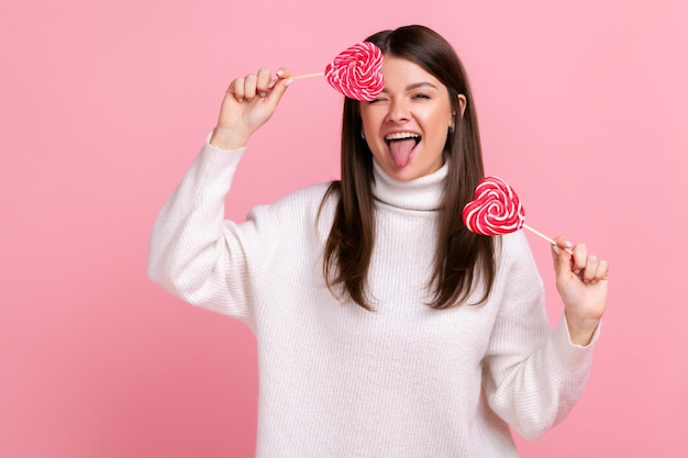 Emocionada mujer feliz cubriendo los ojos con deliciosos dulces en forma de corazón, guiñando un ojo, mostrando la lengua, usando un suéter blanco de estilo casual. Disparo de estudio interior aislado sobre fondo rosa.