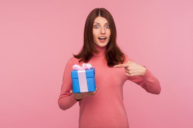 Emocionada mujer morena sorprendida en suéter rosa apuntando con el dedo a la caja de regalo azul en su mano mirando a la cámara con expresión de asombro Disparo de estudio interior aislado sobre fondo rosa