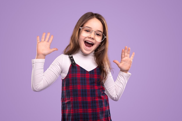 Emocionada niña nerd en uniforme escolar y gafas divirtiéndose y mostrando diez dedos mientras está de pie contra el fondo violeta