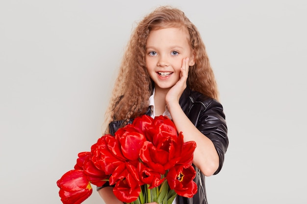 Emocionada niña rubia en chaqueta de cuero negro con ramo de tulipanes rojos, diciendo algo y manteniendo la mano cerca de la boca