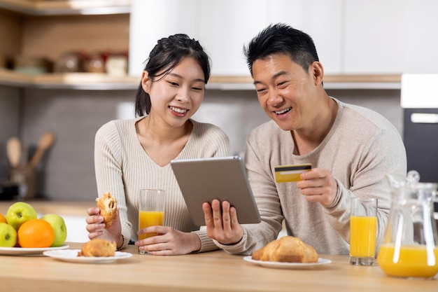 Emocionada pareja asiática comprando desde casa mientras desayuna