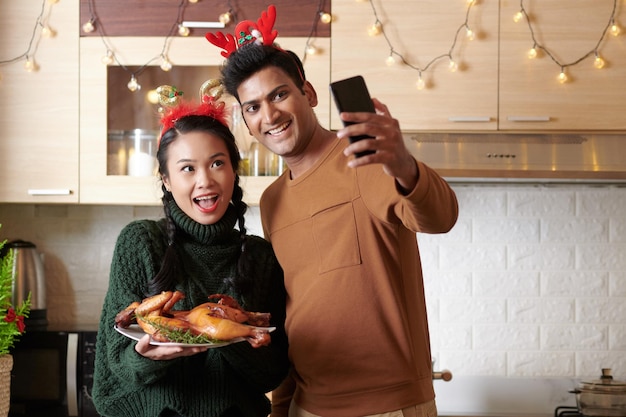 Emocionada pareja joven feliz tomando selfie con pollo frito que cocinaron para la cena de Navidad