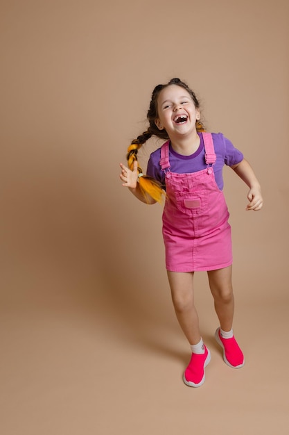 Emocionada, radiante, feliz niña divirtiéndose con dos coletas kanekalon de color amarillo vestidas con mono rosa, camiseta púrpura y zapatillas rosas sobre fondo beige.