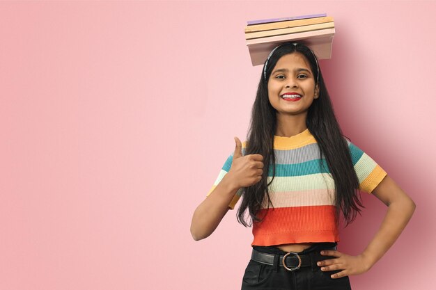 Emocionada y sonriente joven estudiante india asiática posando libros de equilibrio aislados en su cabeza y mostrando los pulgares hacia arriba apuntando a un espacio de copia