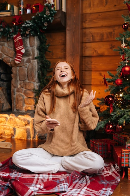 Emocionada sorprendida joven pelirroja vistiendo un suéter de invierno recibiendo buenas noticias en el teléfono móvil