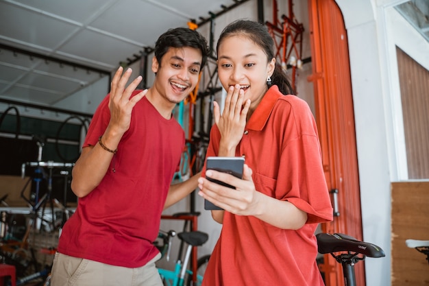 Emocionadas parejas jóvenes asiáticas en la tienda de bicicletas sorprendidas mientras miran el teléfono