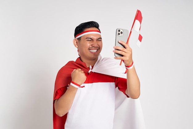 Foto emocionado y apuesto joven asiático con una camiseta blanca casual sosteniendo un teléfono inteligente y una bandera indonesia aislada sobre fondo blanco celebre el día de la independencia de indonesia el 17 de agosto