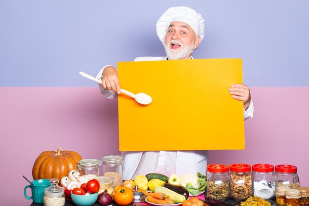 Emocionado chef senior con sombrero de cocinero que señala la cuchara en el menú de la pizarra en blanco Chef divertido que muestra el menú de la pizarra vacía en blanco Chef de cocina