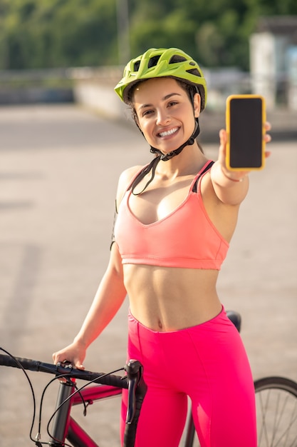 Emocionado. Una chica en ropa deportiva rosa mostrando su teléfono inteligente y mirando emocionada