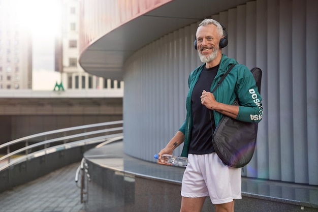 Emocionado deportivo hombre de mediana edad en ropa deportiva y auriculares sonriendo a un lado de pie al aire libre listo