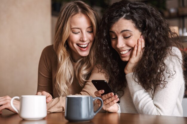 Foto emocionado emocionado sorprendido amigas sentadas en la cafetería con teléfono móvil