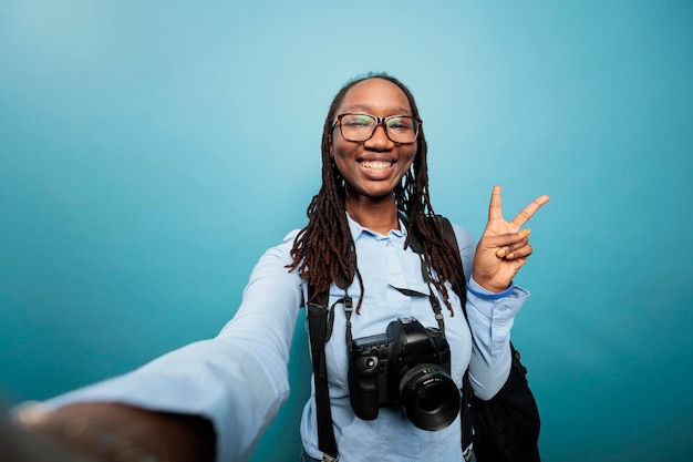 Emocionado entusiasta de la fotografía alegre tomando fotos selfie mientras está de pie sobre fondo azul. Fotógrafo sonriente feliz con cámara DSLR dando señales de paz a la audiencia mientras graba video vlog diario.