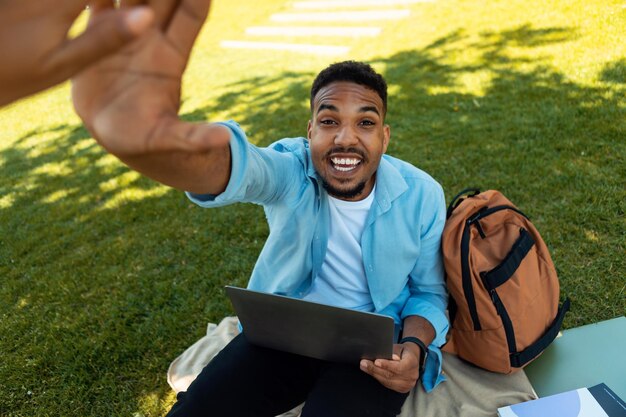 Emocionado estudiante negro dando cinco a un amigo mientras usa una computadora portátil sentada en el césped