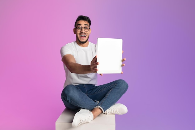 Emocionado hombre árabe milenario mostrando tableta con pantalla en blanco en maqueta de luz de neón para aplicación o sitio web