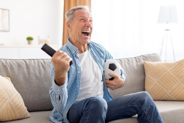 Emocionado hombre maduro viendo la televisión celebrando el gol sosteniendo la pelota