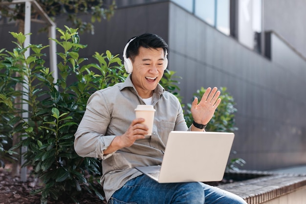 Emocionado hombre de negocios asiático de mediana edad que trabaja en una computadora portátil al aire libre usando auriculares y haciendo videollamadas