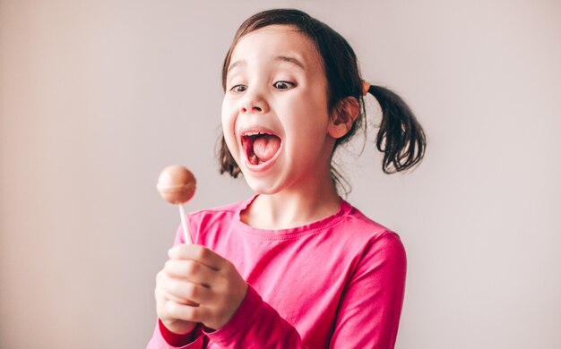 Emocionado increíble joven niña aislada sobre la pared. Mirando a la piruleta con felicidad y placer.