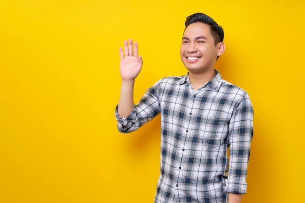 Emocionado joven apuesto hombre asiático vestido con ropa informal Saludando y saludando con una mano mientras se da cuenta de que alguien está aislado en un fondo amarillo Concepto de estilo de vida de la gente