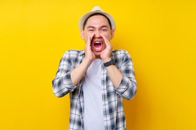 Emocionado joven apuesto hombre étnico asiático de 20 años con ropa informal sombrero gritando compartiendo noticias calientes sobre descuentos de ventas con las manos cerca de la boca mirando la cámara aislada en fondo amarillo