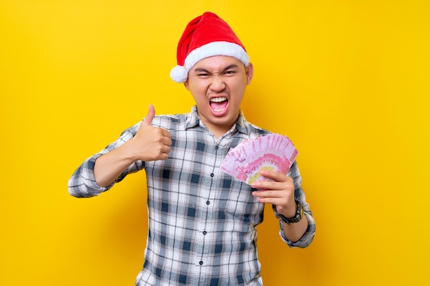 Emocionado joven asiático de 20 años con un sombrero de Navidad mostrando dinero en efectivo rupia y pulgar arriba signo aislado sobre fondo amarillo