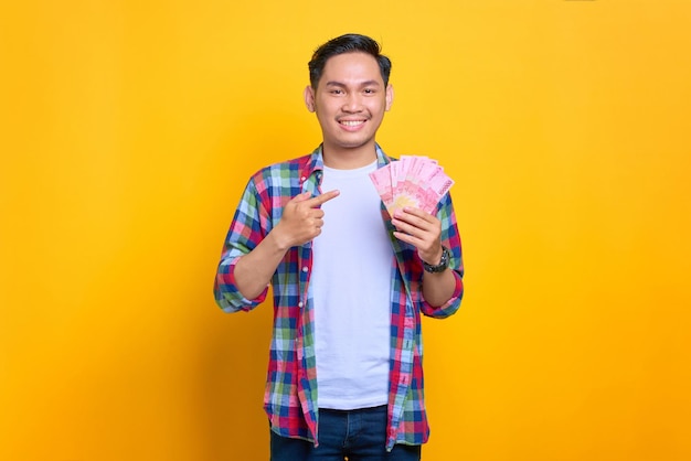 Emocionado joven asiático con camisa a cuadros señalando con el dedo los billetes de dinero aislados de fondo amarillo