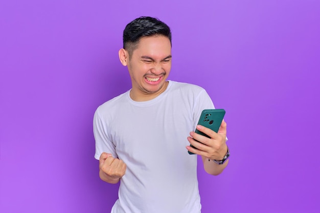 Emocionado joven asiático con camiseta blanca usando un smartphone y haciendo un gesto de ganador aislado en un fondo morado