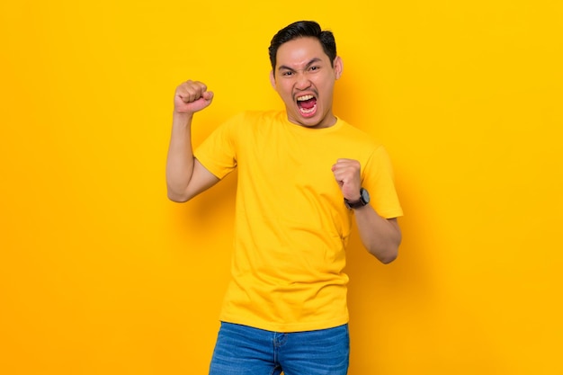 Emocionado joven asiático en camiseta casual celebrando el éxito con el puño levantado aislado sobre fondo amarillo Concepto de estilo de vida de la gente