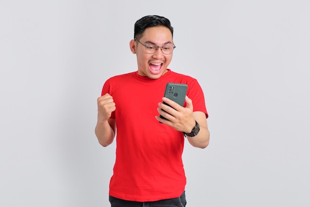 Emocionado joven asiático mirando la pantalla del teléfono móvil haciendo un gesto de sí con el puño levantado reaccionando a las noticias en línea aisladas sobre fondo blanco