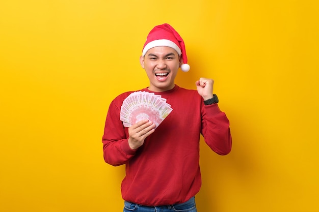 Emocionado joven asiático con sombrero de Papá Noel sosteniendo un abanico de dinero en efectivo y levantando el puño celebrando el éxito en la celebración de fondo de estudio amarillo celebración de Navidad y concepto de Año Nuevo