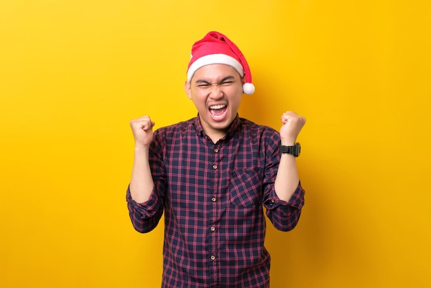 Emocionado joven asiático con sombrero de Santa levantando las manos celebrando el éxito en el fondo amarillo del estudio Feliz Año Nuevo 2023 celebración feliz concepto de vacaciones