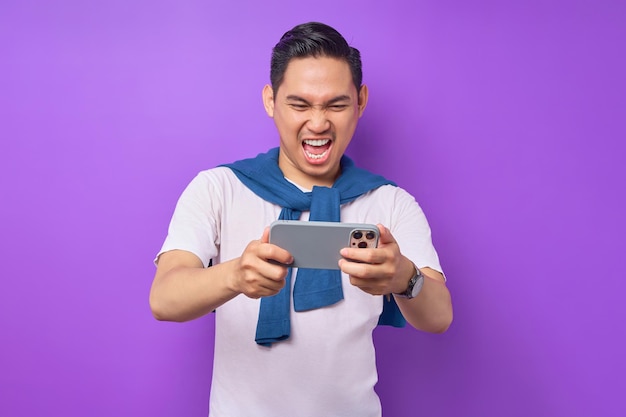 Emocionado joven asiático usando smartphone para jugar juegos móviles aislado sobre fondo púrpura