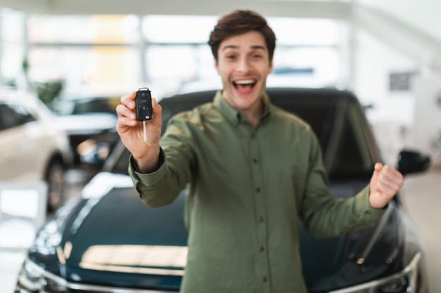 Emocionado joven caucásico mostrando la nueva llave del auto en la cámara gesticulando sí celebrando la compra del auto