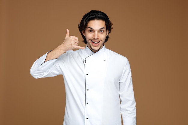 Emocionado joven chef uniformado mirando a la cámara mostrando un gesto de llamada aislado de fondo marrón