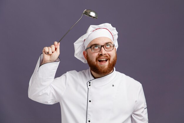 Emocionado joven chef con uniforme de gafas y gorra mirando hacia arriba sosteniendo un cucharón sobre la cabeza aislado en un fondo morado