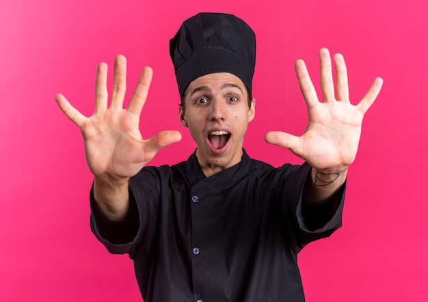 Emocionado joven cocinero de sexo masculino rubio en uniforme de chef y gorra mirando a la cámara mostrando diez con las manos aisladas en la pared rosa