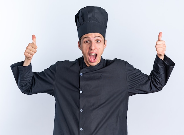 Emocionado joven cocinero de sexo masculino rubio en uniforme de chef y gorra mirando a la cámara mostrando los pulgares para arriba aislado en la pared blanca