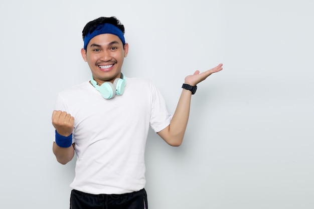 Emocionado joven deportista asiático en diadema azul y camiseta blanca de ropa deportiva con auriculares apuntando con el dedo al espacio de la copia y celebrando el éxito aislado sobre fondo blanco Concepto de deporte de entrenamiento