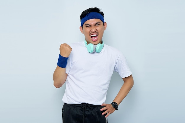 Emocionado joven deportista asiático en diadema azul y camiseta blanca de ropa deportiva con auriculares celebrando la victoria aislado sobre fondo blanco Concepto de deporte de entrenamiento