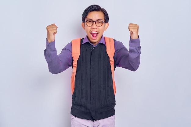 Emocionado joven estudiante asiático de 20 años con ropa informal con mochila mostrando gesto de ganador y celebrando al graduado aislado en fondo gris Concepto de universidad de escuela secundaria